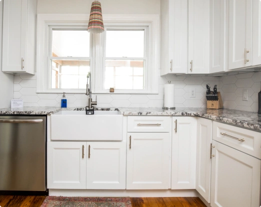 Modern kitchen with white cabinets, farmhouse sink, marble countertop, stainless steel dishwasher, hexagonal tile backsplash, and colorful pendant light.