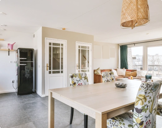 Bright dining and living area with wooden table, floral-upholstered chairs, woven pendant light, glossy black refrigerator, and beige sectional sofa near large windows with green curtains.