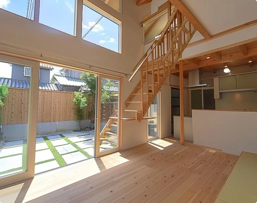 Open-concept home interior with wood flooring, exposed beams, floor-to-ceiling windows overlooking courtyard, and wooden staircase beside modern kitchen.