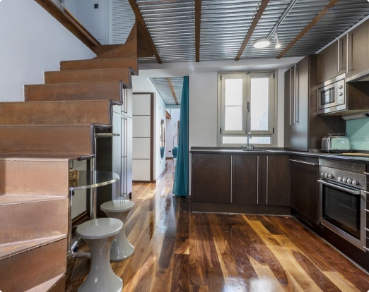 Modern kitchen with dark wood cabinets, stainless steel appliances, polished wood floor, and adjacent staircase with industrial ceiling beams.