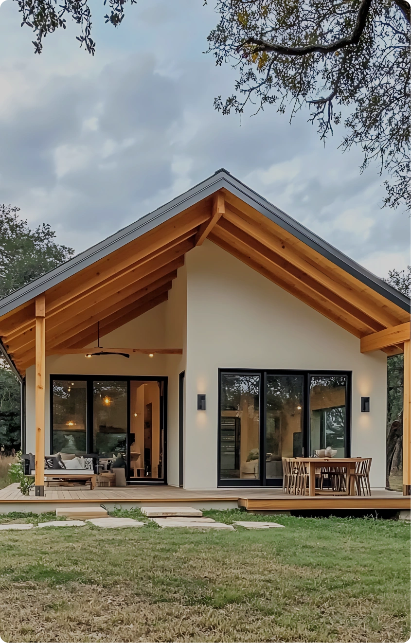 Modern house with steeply pitched roof supported by exposed wooden beams, light-colored exterior walls, large glass sliding doors and windows revealing warmly lit interior with contemporary furnishings, wooden deck with outdoor seating, grassy yard with stone pavers, and trees under cloudy sky.
