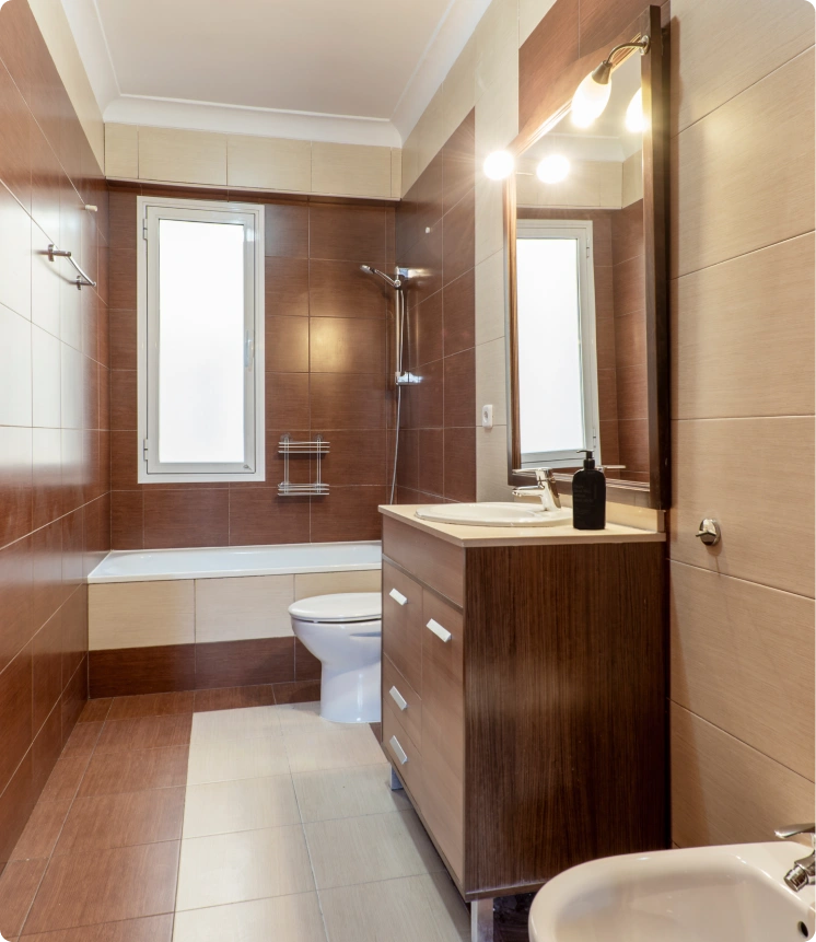 A modern bathroom with brown and beige tiled walls, a bathtub with shower fixtures, a toilet, a wooden vanity with a sink and mirror, and a narrow frosted window.