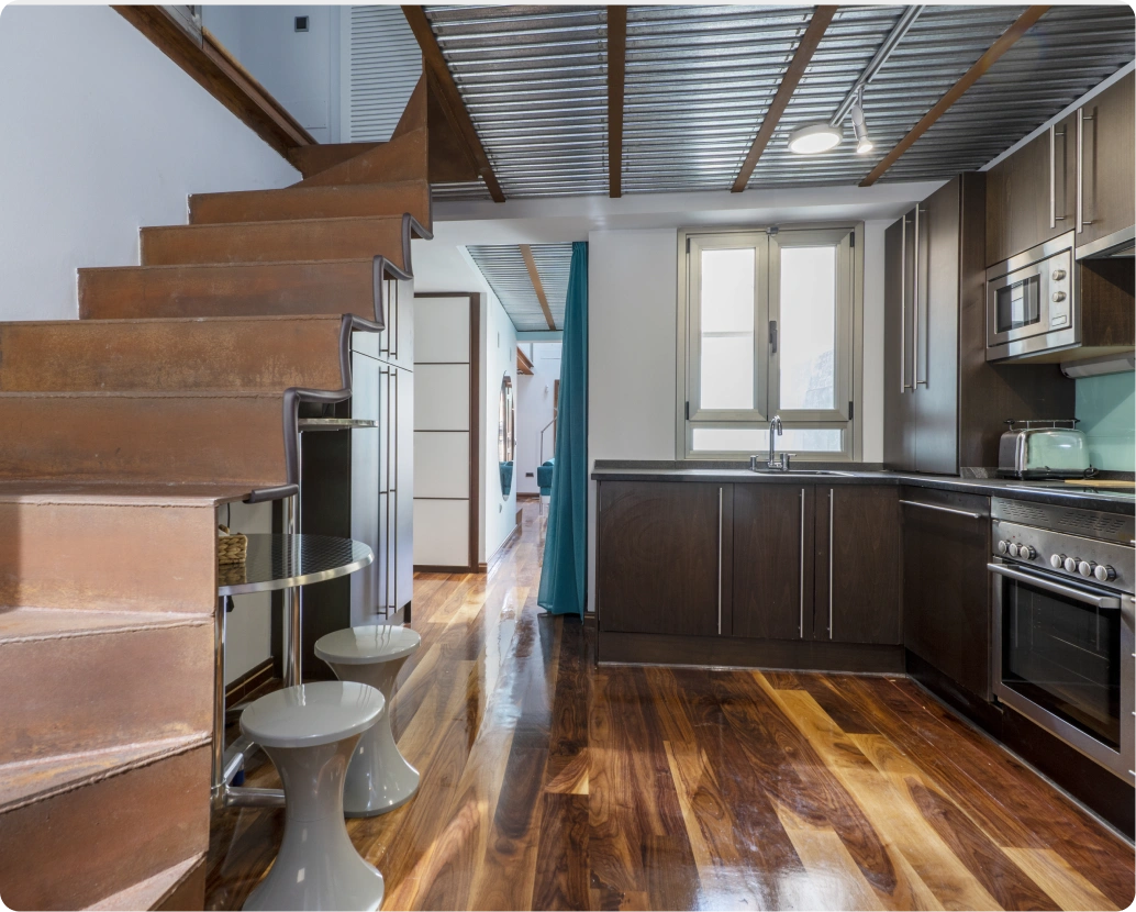 Modern interior with rust-colored metal staircase, round table with two white stools beneath, polished wood floor, kitchen with dark cabinets and stainless steel appliances, and corrugated metal ceiling with wooden beams.