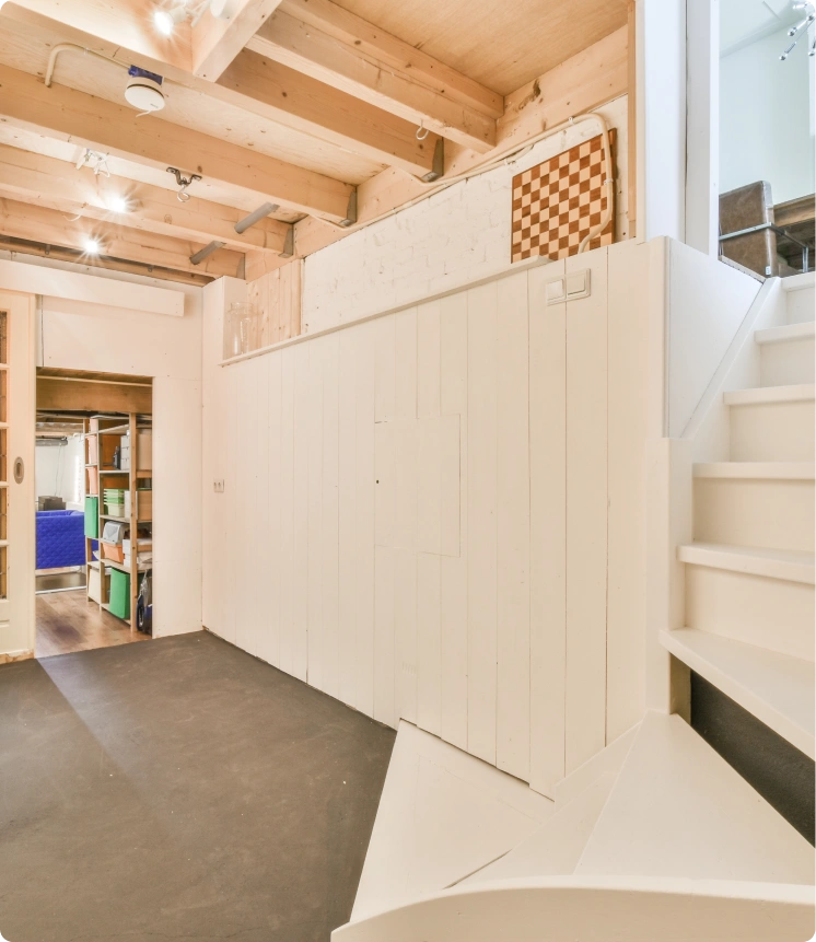 Minimalist interior with light wood and white surfaces, staircase leading to upper level with checkerboard panel, exposed wooden beams with track lighting, dark polished floor, and doorway to room with shelves and blue furniture.