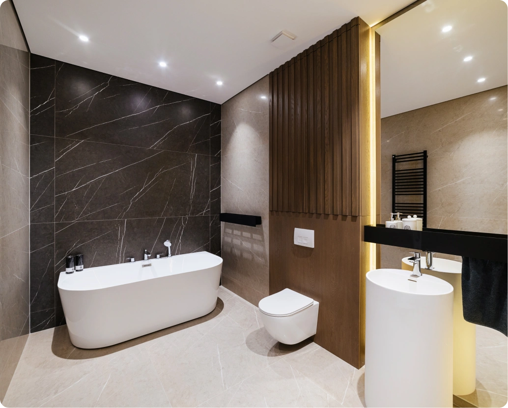 Luxury bathroom remodeling featuring a freestanding white soaking tub against a dramatic black marble accent wall, a wall-hung toilet for easy cleaning, a modern pedestal sink, and warm wood slat paneling with integrated LED lighting.