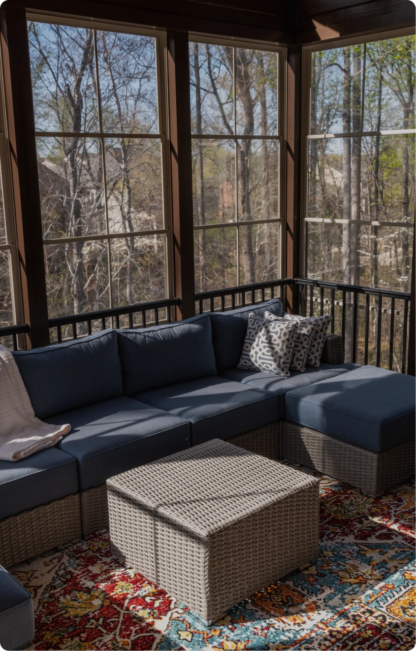 Sunroom home addition featuring a spacious L-shaped outdoor sectional with navy blue cushions, a woven wicker storage ottoman, and a vibrant patterned rug. Floor-to-ceiling windows offer panoramic views of the surrounding wooded landscape and backyard.