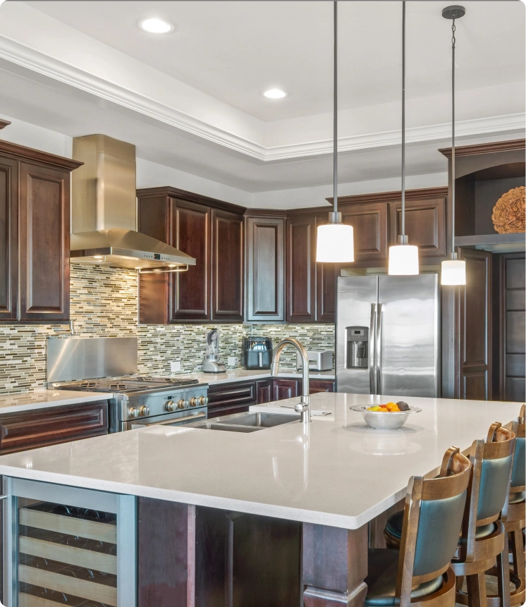 Luxury kitchen remodel featuring rich dark wood cabinetry, a large central island with a white quartz countertop and built-in wine refrigerator, stainless steel appliances including a professional-grade range and hood, a mosaic tile backsplash, and elegant pendant lighting over the dining area.