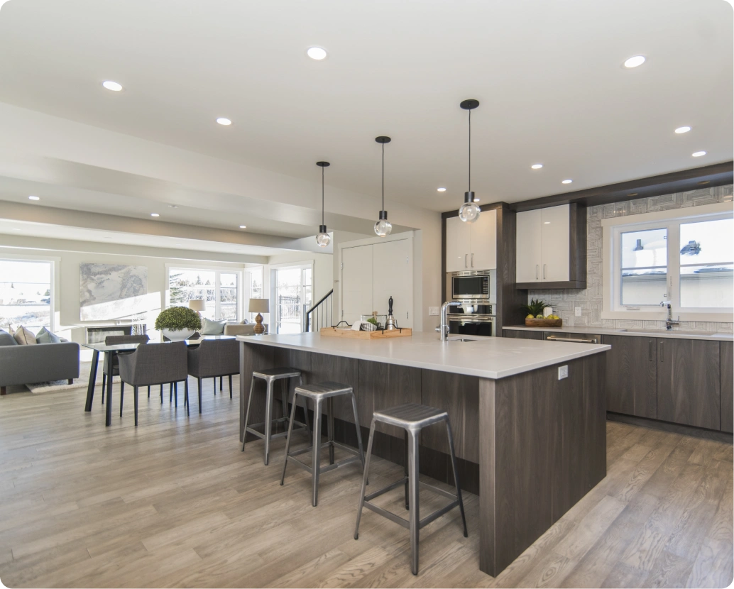 Contemporary open-concept kitchen and living area featuring a large central island with dark wood cabinetry, a light quartz countertop, and three metal bar stools. The space includes two-tone cabinetry, pendant lighting, recessed ceiling lights, light wood-look flooring, and a seamless flow into a bright dining and living area with large windows and modern furnishings.